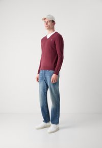 Pulover bordeaux cu guler în V dintr-o țesătură moale, purtat peste un tricou alb, asortat cu blugi din denim albastru deschis și adidași albi.