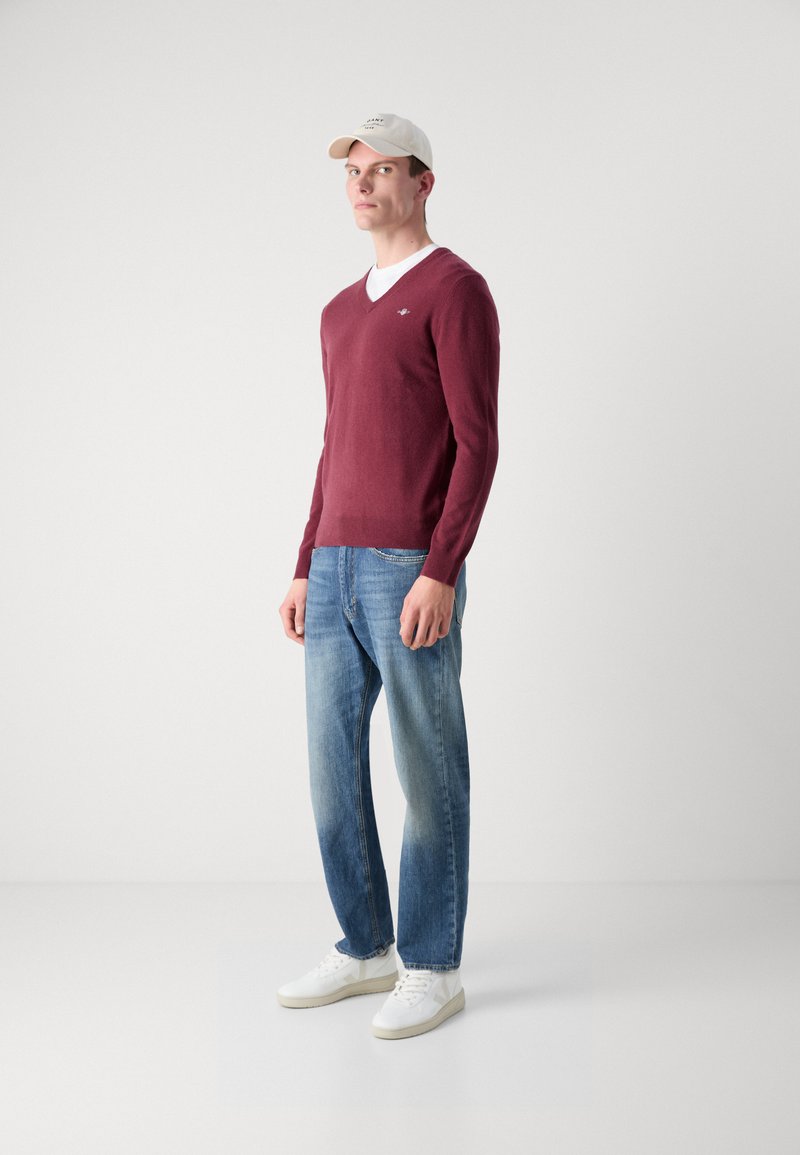 Suéter burdeos de cuello en V en tejido suave, llevado sobre una camiseta blanca, combinado con jeans de mezclilla azul claro y zapatillas blancas.