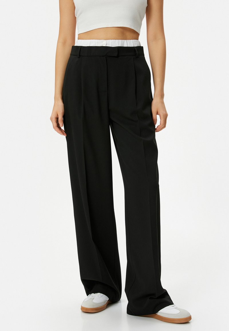 Zwarte wijde broek met een getailleerde pasvorm, voorzien van een knoopsluiting en een gestructureerde tailleband. Gecombineerd met witte sneakers.