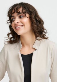 Beige lätt jacka med mandarin-krage, slät textur; bärs över en svart ribbad topp, vilket framhäver en neutral färgpalett.