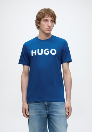Ung mand iført en blå T-shirt med "HUGO" i hvide bogstaver, kombineret med lyseblå jeans, stående mod en ensfarvet lys baggrund.
