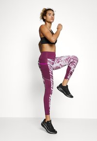 Brassiére sportivo nera, leggings con motivo viola e bianco e tasche laterali, e scarpe da ginnastica nere. Modello in una posa attiva.