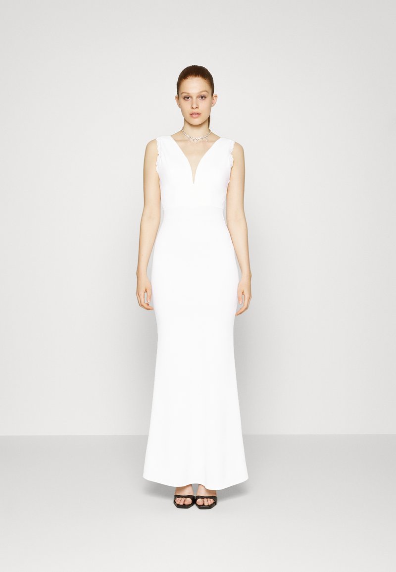 Vestido branco sem mangas com decote em V e parte inferior ampla, apresentando bordas rendadas nos ombros. Modelo usa sandálias pretas com salto bloco.