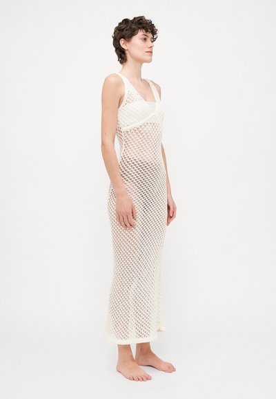 Vêtement : robe longue sans manches en tricot ouvert faite de fil blanc ; présente un corsage ajusté et une jupe ample et fluide avec un design à longueur complète.