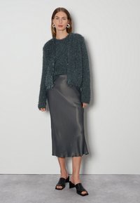 Türkisfarbener, fellig weicher Cardigan über einem passenden, felligen Pullover, kombiniert mit einem knielangen, schwarzen Satinrock und schwarzen, offenen Mules. Durchgehend glatte Textur.