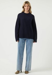 Marineblauwe gebreide trui met een ronde hals, ruime pasvorm en verlaagde schouders, gecombineerd met lichtblauwe wijde jeans en bruine enkellaarzen met hak.