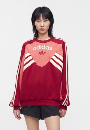 Młoda osoba z czarnymi włosami nosi czerwoną i różową bluzę Adidas oraz czarne spodenki, stoi na jednolitym jasnym tle.