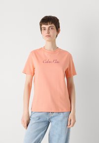 Šviesiai persikinis medvilninis marškinėliai su apvaliomis apykaklėmis, trumpomis rankovėmis ir išsiuvinėtu "Calvin Klein" logotipu tamsesniu atspalviu. Dėvimi su šviesiai mėlynomis džinsais.