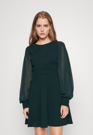 Dunkelgrünes Kleid mit tailliertem Oberteil und weit schwingendem Rock. Verfügt über transparente Puffärmel und eine glatte Oberfläche. Einfacher runder Ausschnitt.