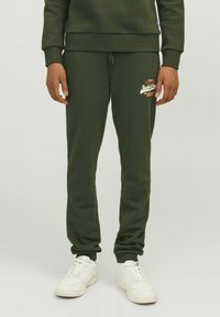 Olivengrønne bomulds sweatpants med en tapered design, ribbede manchetter og et logo trykt på venstre lår. Sammen med hvide sneakers.