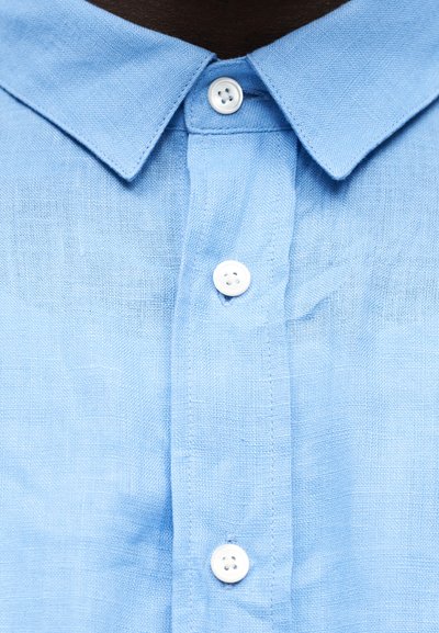 Γιακάς πουκαμίσου από ανοιχτό γαλάζιο λινό ύφασμα με τρία λευκά κουμπιά κουμπωμένα, φορεμένος από άτομο με σκουρόχρωμο δέρμα.