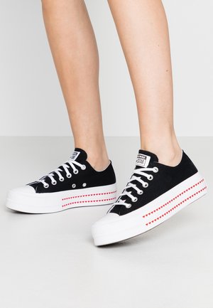 Jambes portant des baskets basses noires avec des lacets et des semelles blanches décorées de petits motifs de cœurs rouges le long de la semelle intermédiaire.