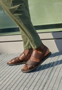 Braune Leder-Sandalen mit einem Riemendesign und einem Schnallendetail, kombiniert mit grünen Hosen auf einer strukturierten Oberfläche.