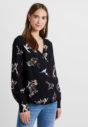 Jeune femme aux longs cheveux bruns, portant un blouse noire à motifs floraux et d'oiseaux, et un jean bleu, se tenant devant un fond clair.