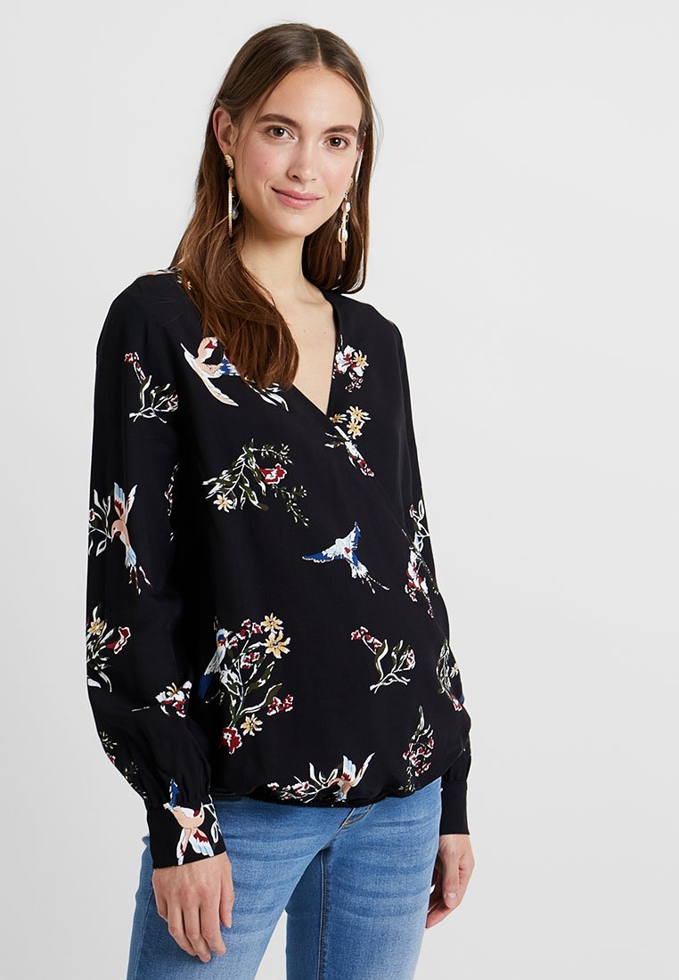 Jeune femme aux longs cheveux bruns, portant un blouse noire à motifs floraux et d'oiseaux, et un jean bleu, se tenant devant un fond clair.