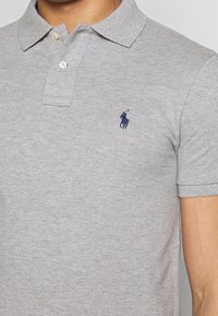 Närbild av en person som bär en ljusgrå polo med en liten mörkblå broderad logotyp på bröstet.
