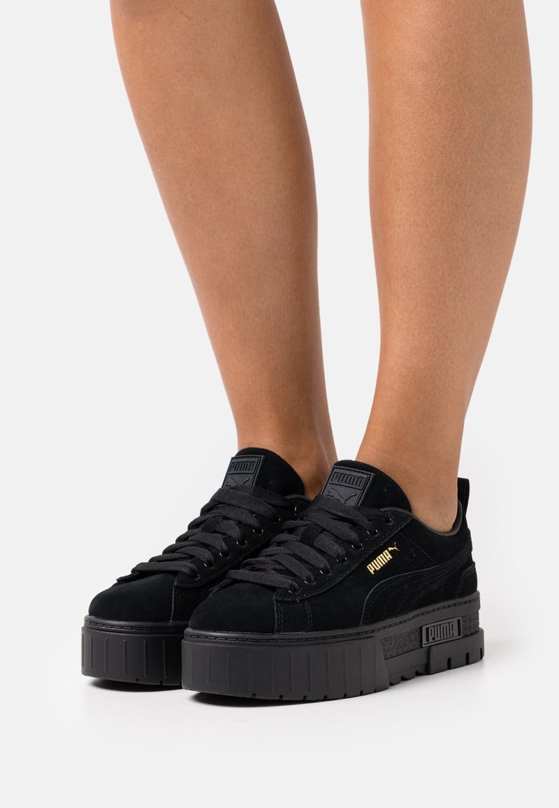 Jambes portant des baskets à plateforme Puma noires avec semelles épaisses, texture suédée, style à lacets et logo doré Puma sur les côtés.