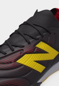 Κοντινό πλάνο μαύρου αθλητικού παπουτσιού με κόκκινες τελείες, μαύρα κορδόνια και μεγάλο κίτρινο ανάγλυφο λογότυπο στο πλάι.