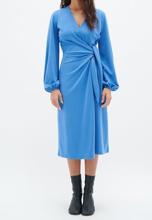 Donna che indossa un vestito azzurro avvolgente a maniche lunghe, lungo fino al ginocchio, con dettaglio di vita arricciata e stivaletti neri alla caviglia, in piedi davanti a uno sfondo bianco.