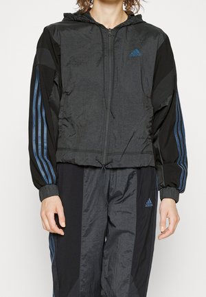 Personne portant un survêtement Adidas noir avec des bandes bleues, une veste à fermeture éclair avec capuche, et un pantalon assorti, debout les bras détendus.