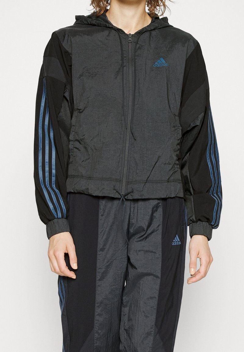 Personne portant un survêtement Adidas noir avec des bandes bleues, une veste à fermeture éclair avec capuche, et un pantalon assorti, debout les bras détendus.