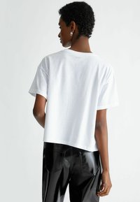 T-shirt bianca in cotone con design cropped e maniche corte. Presenta un piccolo logo vicino allo scollo. Abbinata a pantaloni neri lucidi.