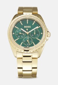Montre dorée avec un cadran vert, accents en cristal, trois sous-cadrans et un bracelet en or poli. Présente une lunette texturée et des indicateurs de date.