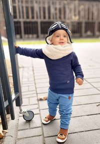 Niebieski bawełniany sweter, jasnoniebieskie dresy, beżowy polarowy szalik, buty z twarzą brązowego misia i czapka pilotka z goglami. Trzyma bramę.
