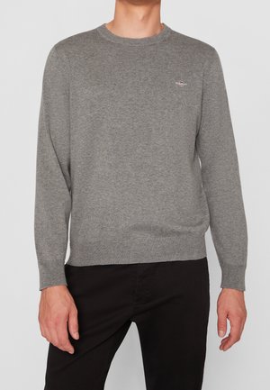 Homme portant un pull en maille gris à manches longues avec un petit logo sur la poitrine, associé à un pantalon noir, sur un fond uni.
