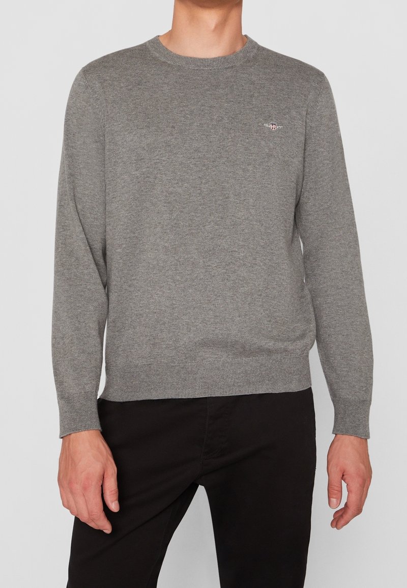 Homme portant un pull en maille gris à manches longues avec un petit logo sur la poitrine, associé à un pantalon noir, sur un fond uni.