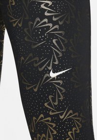 Černé legíny s metalicky zlatým květinovým vzorem a puntíkovými akcenty, se bílým logem Nike v levém dolním rohu.