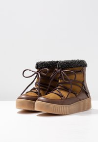 Botas de piel marrón hasta el tobillo con diseño de cordones, que presentan un cuerpo de ante color canela, un collar negro suave y una suela de goma texturizada.