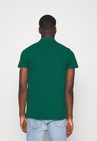 Homme portant un polo à manches courtes vert et un jean bleu clair, de dos sur un fond blanc uni.