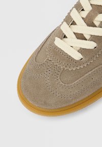 Biqueira de ténis em camurça bege com atacadores creme e costuras detalhadas, apresentando uma borda da sola em borracha texturada castanho claro.