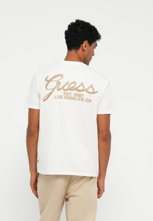 Camiseta blanca de algodón con un gran logo beige de "Guess" en la parte posterior, escrito en cursiva con "EST. 1981 LOS ÁNGELES, CA" debajo. Mangas cortas.
