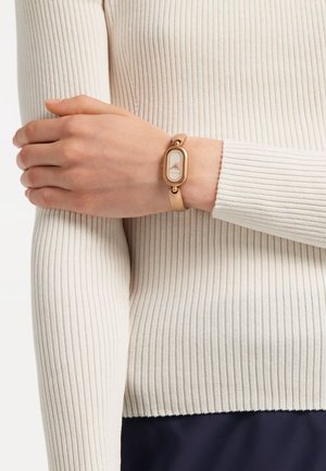 Bracciale in pelle di un marrone chiaro con un quadrante ovale in tonalità oro; presenta un quadrante bianco con un motivo testurizzato; indossato su una manica di maglione a coste.