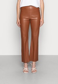 Vrouw in bruine faux leren cropped broeken en witte sandaaltjes met hoge hakken, staande tegen een effen lichte achtergrond.