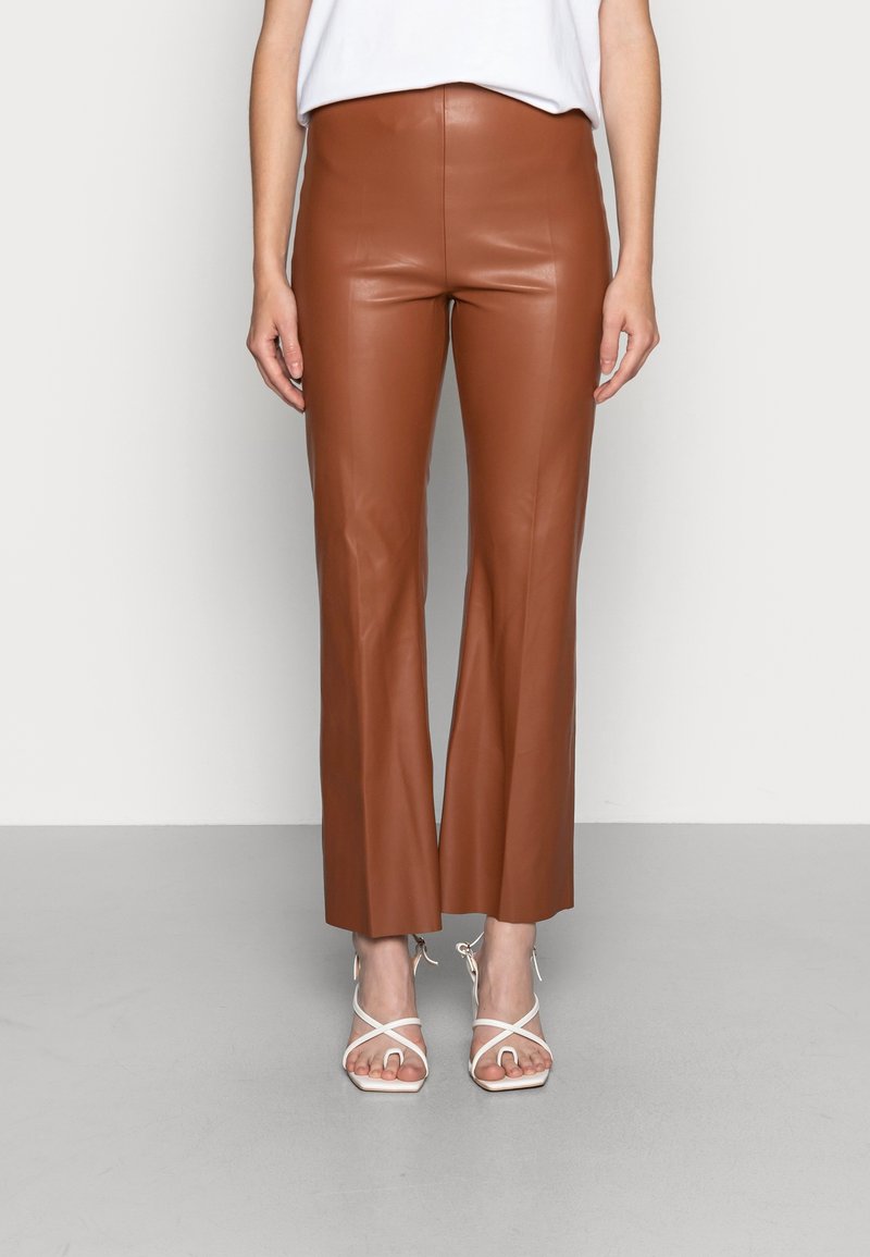 Vrouw in bruine faux leren cropped broeken en witte sandaaltjes met hoge hakken, staande tegen een effen lichte achtergrond.