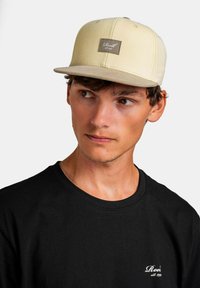 Beige flat-brimmed cap with a stitched brown label on the front, worn with a black short-sleeve shirt featuring white text.