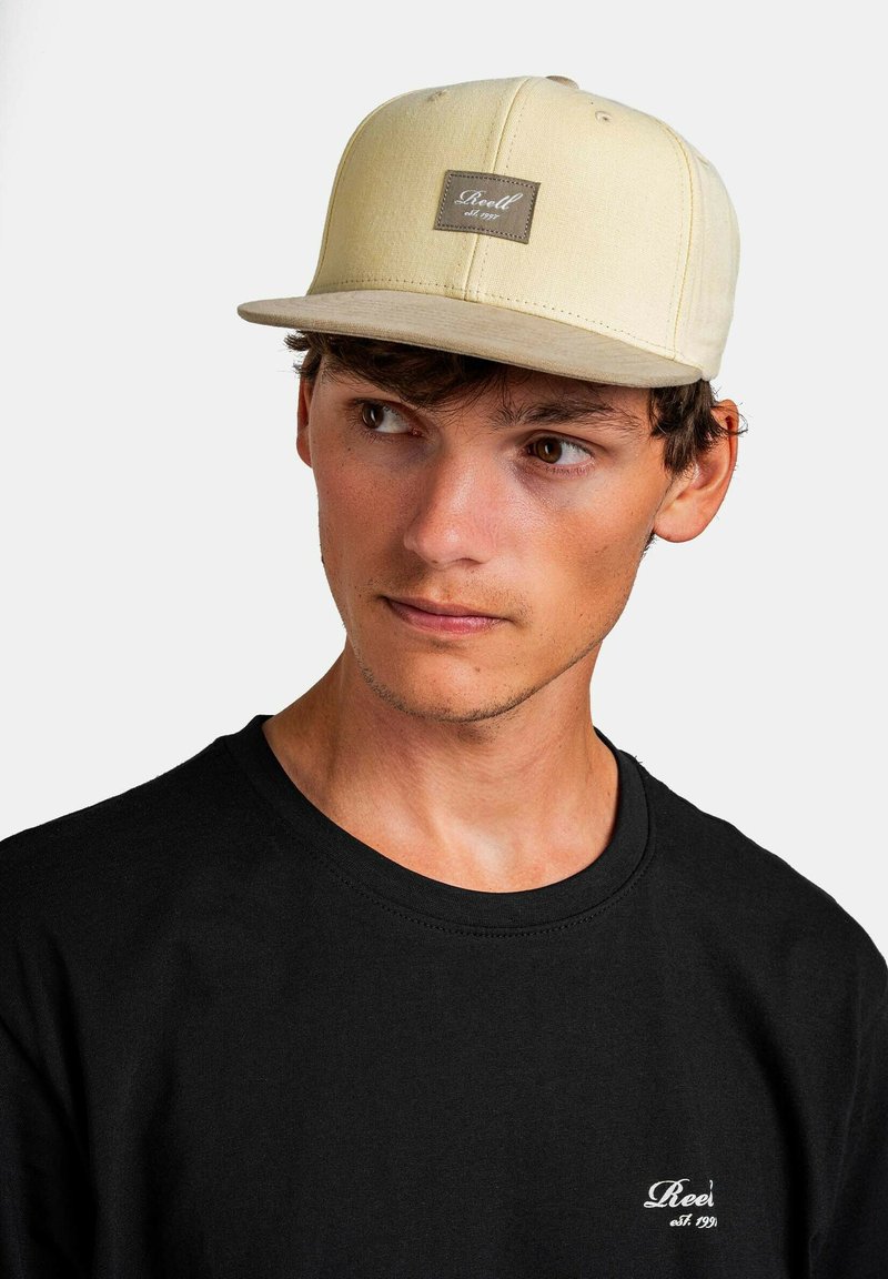 Beige flat-brimmed cap with a stitched brown label on the front, worn with a black short-sleeve shirt featuring white text.