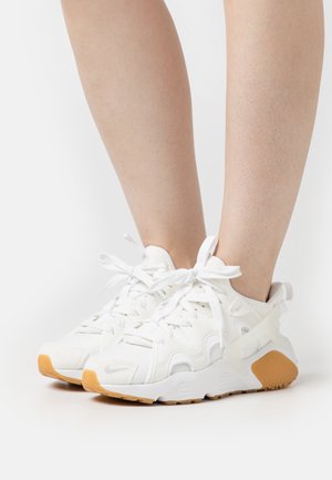 Baskets blanches de sport avec semelles en caoutchouc couleur beige portées sur des jambes nues, lacées avec des lacets blancs noués, sur un fond blanc uni.