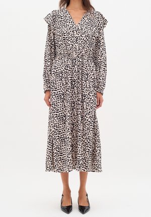 Donna con un lungo vestito leopardato con scollo a V, maniche lunghe, vita elastica e tacchi neri a punta, in piedi contro uno sfondo bianco.