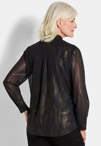 Schwarze transparente Bluse mit langen Ärmeln und Knopfmanschetten, die ein dezentes Muster und einen Rückenfalten für Bewegungsfreiheit aufweist.
