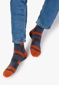 Mehrfarbige Socken in Lila, Orange, Grün und Schwarz, mit einem geometrischen Muster und kontrastierenden Zehenakzenten, getragen mit blauen Jeans.