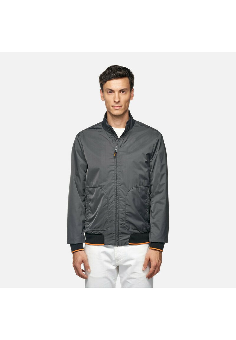 Giacca bomber grigia con zip frontale, due tasche anteriori e polsini a coste con dettagli arancioni. Realizzata in tessuto leggero e liscio.