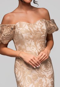 Off-shoulder klänning i genomskinlig beige med blommönster. Mjuka puffärmar, figursydd bodice och en delikat armlänk på händerna. Slät textur.