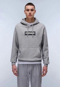 Giovane uomo che indossa un hoodie grigio chiaro con tasca frontale e logo Napapijri, abbinato a pantaloni grigi, in piedi contro uno sfondo neutro.