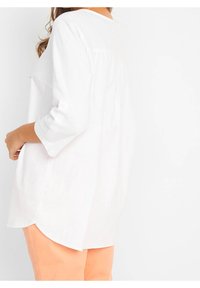 Haut blanc, ample, avec un ourlet arrondi et des manches trois-quarts, doté d'un détail à boutons et d'un tissu texturé. Porté avec un pantalon orange clair.