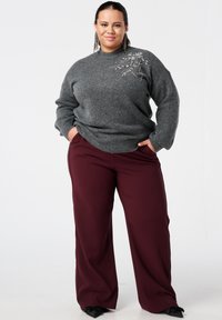 Grijze trui met een gestructureerde afwerking en versieringen op de schouder, gecombineerd met bordeauxrode wijde broekspijpen en zwarte schoenen.