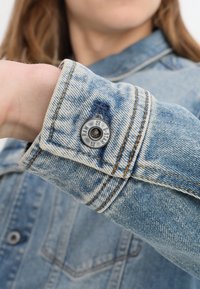 Ljusblå denimjacka med ärm och en metallknapp märkt "G-STAR RAW," delvis vy av en person som bär den.